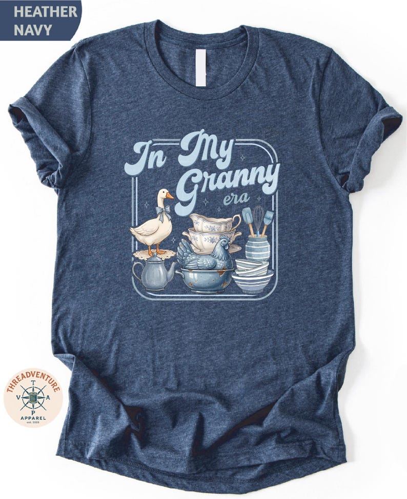 A navy blue t-shirt with a graphic design depicting a white duck surrounded by various kitchen items, including a teapot, bowls, and spoons.