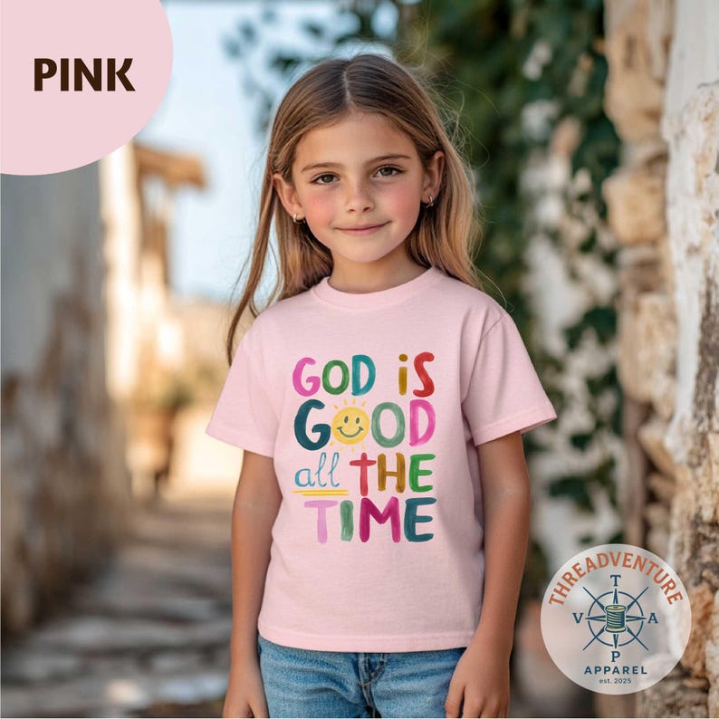 May include: A light pink t-shirt with the words "GOD IS GOOD all THE TIME" in colorful letters and a smiling sun graphic. The shirt is worn by a young person. The Threadventure Apparel logo is visible in the bottom right corner.