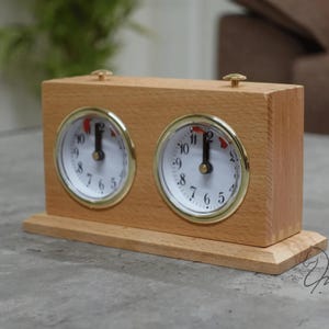 Könnte beinhalten: Eine rechteckige, helle analoge Spieluhr aus Holz mit zwei runden Zifferblättern, goldfarbenen Ringen, weißem Hintergrund, schwarzen Ziffern und schwarzen Zeigern. Auf der Oberseite des Holzgehäuses befinden sich zwei kleine messingfarbene Knöpfe.