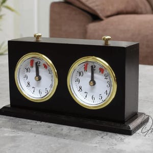 Può includere: Un orologio da gioco analogico rettangolare in legno nero con due quadranti circolari a sfondo bianco, numeri neri e cornici dorate. Due piccoli pulsanti dorati sono posizionati sulla parte superiore dell'alloggiamento nero per l'uso manuale.