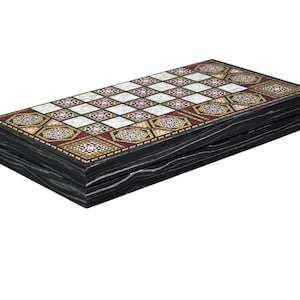 Op de afbeelding: Een rechthoekig backgammonbord met een zwarte houtnerf buitenkant. Het speeloppervlak toont een raster van witte vierkanten afgewisseld met ingewikkelde geometrische patronen in rood, goud, wit en zwart, omrand door een decoratief kader.