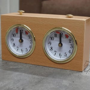 Op de afbeelding: Een rechthoekige analoge schaakklok van lichtgekleurd hout met twee ronde wijzerplaten, witte achtergronden, zwarte cijfers, goudkleurige metalen randen en kleine rode vlaggetjes op de twaalf-uur-positie, bekroond met twee goudkleurige knoppen.