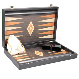 May include: An open wooden backgammon board featuring a dark brown exterior and a playing surface with alternating light and dark brown triangular points. The board includes two black dice cups, a cream-coloured drawstring bag, and the text Staunton.
