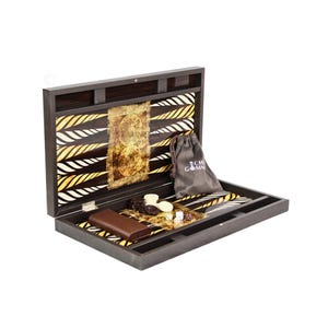 May include: A dark wooden backgammon set featuring a hinged board with a gold and black triangular point pattern. The set includes a brown leather dice cup, a grey drawstring pouch with white text, white and dark brown checkers, and two dice.