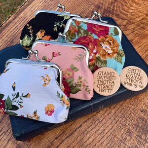 May include: Four small floral patterned coin purses with silver clasps, sitting on top of a black leather-bound book with two wooden circular tags with text. The text on the tags reads "GTAMD OATNBTI TNOOTFA OTSAOOT HSTTTT" and "OATTI HOYAL IAWYAE TTEOTA M28:19-20".