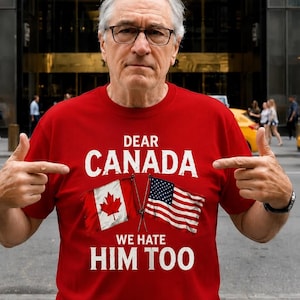 May include: A red short-sleeved t-shirt featuring the white text DEAR CANADA WE HATE HIM TOO and a graphic of a Canadian flag and an American flag crossed in the center. The shirt is made of a solid red fabric with a crew neckline and standard fit.