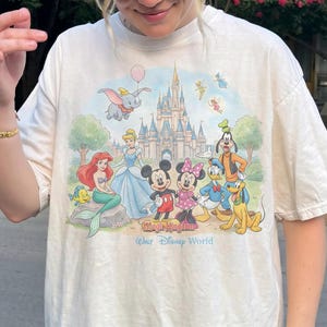 May include: Cream-colored t-shirt featuring a colorful graphic of Disney characters including Mickey Mouse, Minnie Mouse, Donald Duck, and Ariel. The graphic also includes Cinderella's castle and the text "Magic Kingdom Walt Disney World".