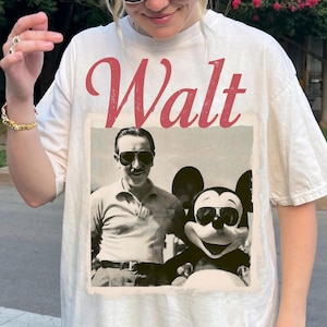 May include: Cream-coloured t-shirt with a vintage graphic of Walt Disney and Mickey Mouse in sunglasses. The word "Walt" is printed in red script. The shirt has a relaxed fit and is made of a soft material.