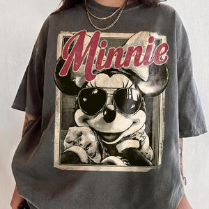 May include: Gray oversized t-shirt featuring a vintage graphic of Minnie Mouse wearing sunglasses. The word "Minnie" is printed in red script above the image. The shirt has short sleeves and a relaxed fit.