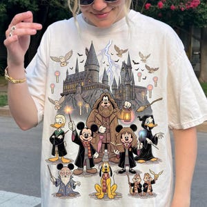 May include: Cream-colored t-shirt featuring a whimsical illustration of a castle, owls, lightning bolts, and various cartoon characters dressed in wizard attire. The characters include Mickey Mouse, Minnie Mouse, Donald Duck, and others. The design is a playful blend of fantasy and animation.
