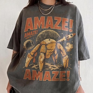 May include: Dark gray t-shirt with a retro space-themed graphic. The design features a crab-like creature, a rocket ship, and the word "AMAZE!" in orange. An astronaut helmet and a planet are also included in the design.