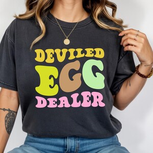 May include: Dark gray t-shirt with the words "DEVILED EGG DEALER" in yellow, green, brown, and pink. The text has a retro, wavy font. The shirt is a casual, unisex style.