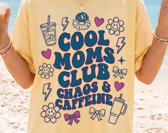 May include: Yellow t-shirt with the text "COOL MOMS CLUB CHAOS & CAFFEINE" in blue and pink. The design includes illustrations of a coffee cup, hearts, flowers, bows, and lightning bolts. The shirt is a light yellow color.