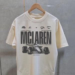 May include: Cream-colored t-shirt featuring a black graphic of a Formula One race car and the word "MCLAREN" in bold letters. The text "LANDO NORRIS" and "OSCAR PIASTRI" are above the word "MCLAREN". The words "FORMULA ONE TEAM" and "TEAM PAPAYA" are also printed on the shirt.