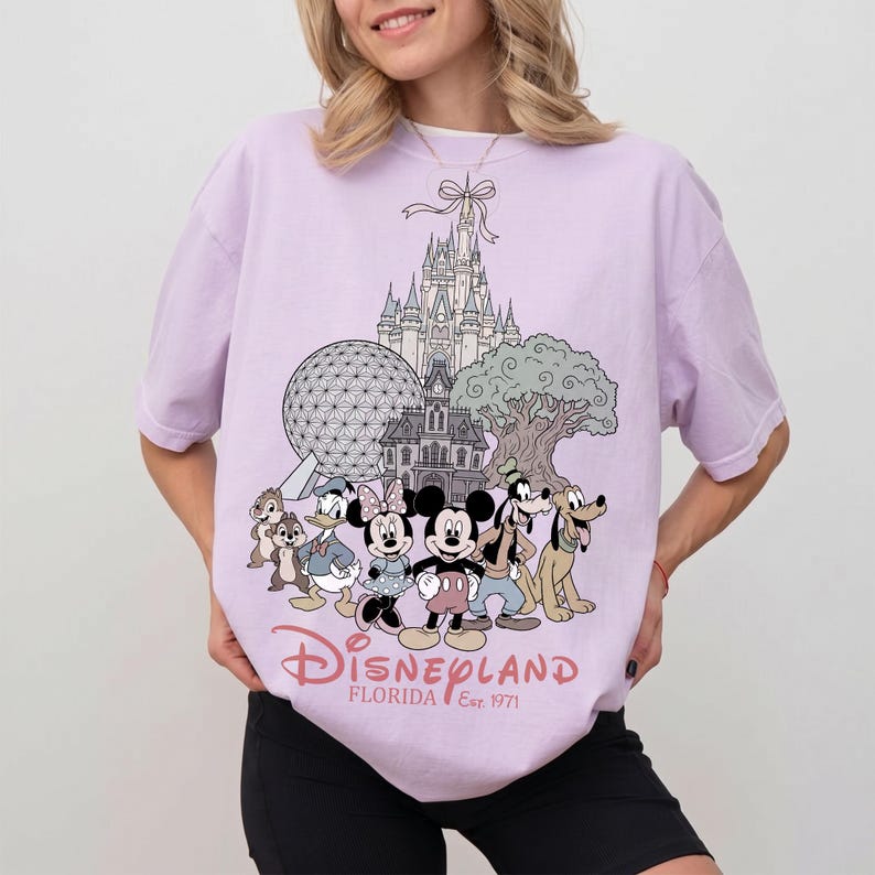K&ouml;nnte beinhalten: Helllila T-Shirt mit einem Disneyland Florida-Motiv. Das Design zeigt Micky und Minnie Maus, Donald Duck, Goofy und Pluto sowie Cinderella's Schloss, Spaceship Earth und den Tree of Life.