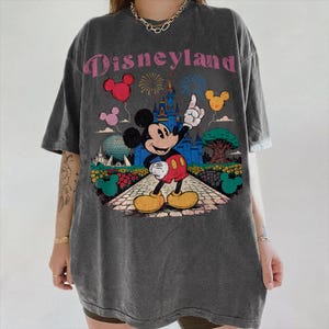 Puede incluir: Camiseta gris oscuro con un dise&ntilde;o de Disneyland. Mickey Mouse se&ntilde;ala un castillo, con globos, fuegos artificiales y un &aacute;rbol en el dise&ntilde;o. El texto "Disneyland" es rosa.