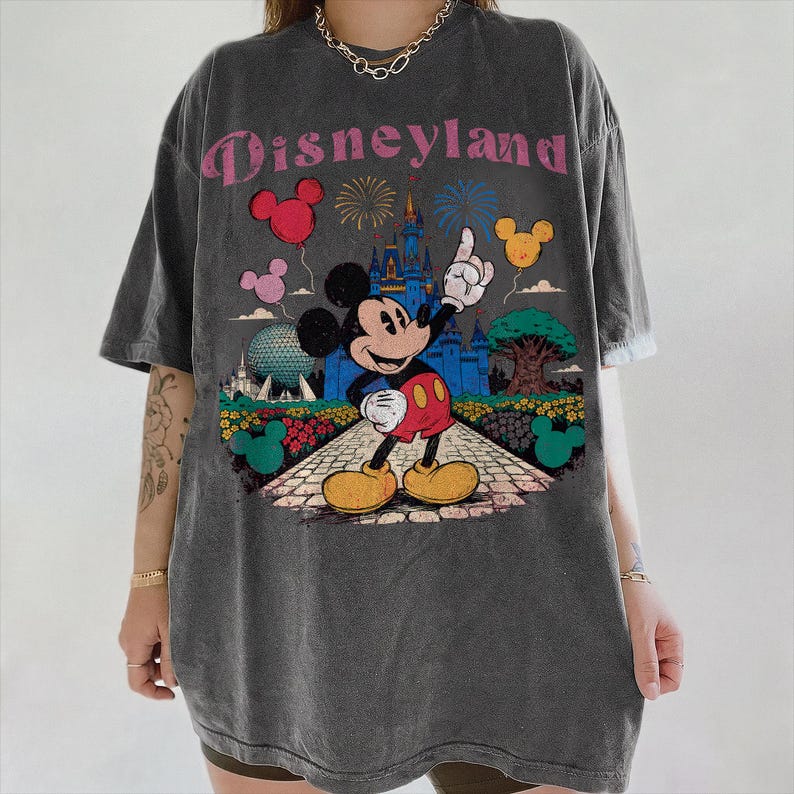 Puede incluir: Camiseta gris oscuro con un dise&ntilde;o de Disneyland. Mickey Mouse se&ntilde;ala un castillo, con globos, fuegos artificiales y un &aacute;rbol en el dise&ntilde;o. El texto "Disneyland" es rosa.