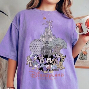 K&ouml;nnte beinhalten: Ein hellviolettes T-Shirt mit einem Disneyland-Motiv. Das Design zeigt Micky Maus, Minnie Maus und andere Charaktere sowie ikonische Wahrzeichen. Der Text lautet "Disneyland Est. 1971".