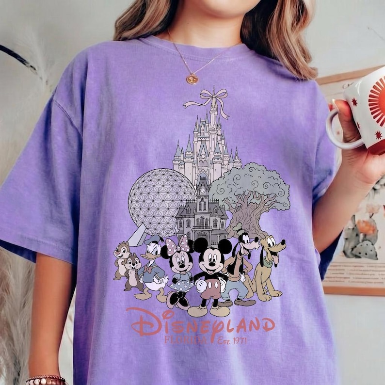 K&ouml;nnte beinhalten: Ein hellviolettes T-Shirt mit einem Disneyland-Motiv. Das Design zeigt Micky Maus, Minnie Maus und andere Charaktere sowie ikonische Wahrzeichen. Der Text lautet "Disneyland Est. 1971".
