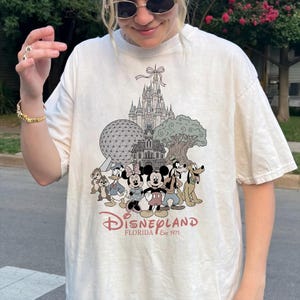 K&ouml;nnte beinhalten: Cremefarbenes T-Shirt mit einem Disneyland Florida-Motiv. Das Design zeigt ein Schloss, Charaktere wie Mickey und Minnie Mouse und den Text "Disneyland Florida Est. 1971". Das Shirt hat eine lockere Passform.