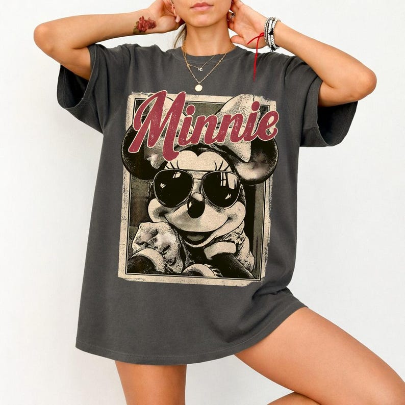 May include: Dark gray t-shirt featuring a vintage-style graphic of Minnie Mouse wearing sunglasses. The word "Minnie" is printed in a red, cursive font above the image. The shirt has short sleeves and a relaxed fit.