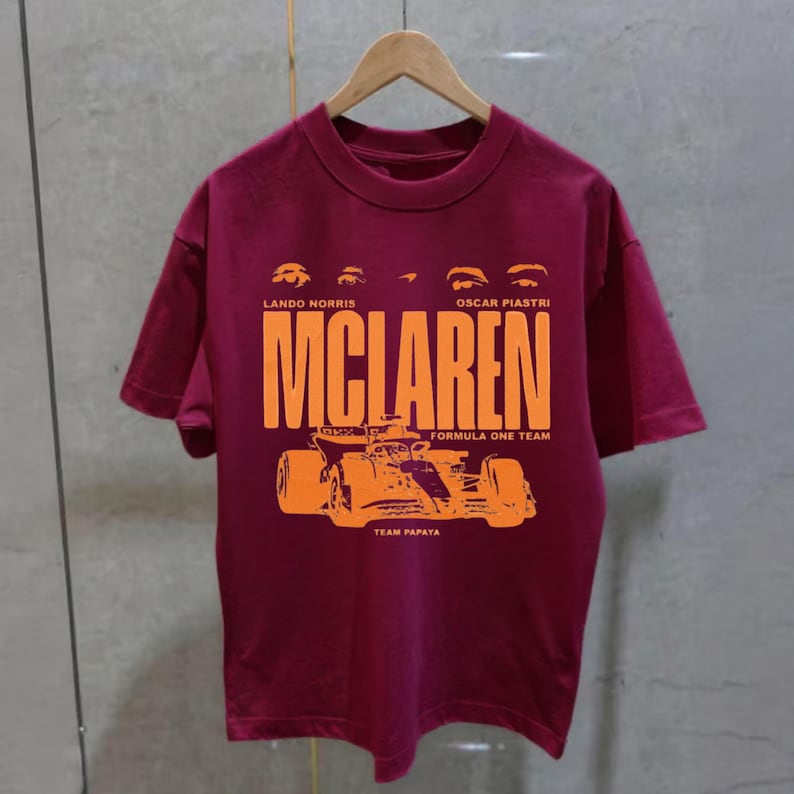 May include: Burgundy t-shirt featuring the McLaren Formula One Team logo in orange. The design includes the names Lando Norris and Oscar Piastri, along with a graphic of a race car. The shirt is hanging on a wooden hanger.