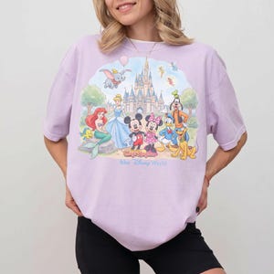 May include: A light purple t-shirt featuring a colorful graphic of Disney characters including Mickey Mouse, Minnie Mouse, Donald Duck, and Cinderella. The text "Magic Kingdom" and "Walt Disney World" are also visible.