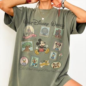 May include: Olive green t-shirt featuring a Walt Disney World design. The shirt showcases various Disney characters and park icons within postage stamp-like frames. Text on the shirt reads "Walt Disney World" and "The Most Magical Place On Earth."