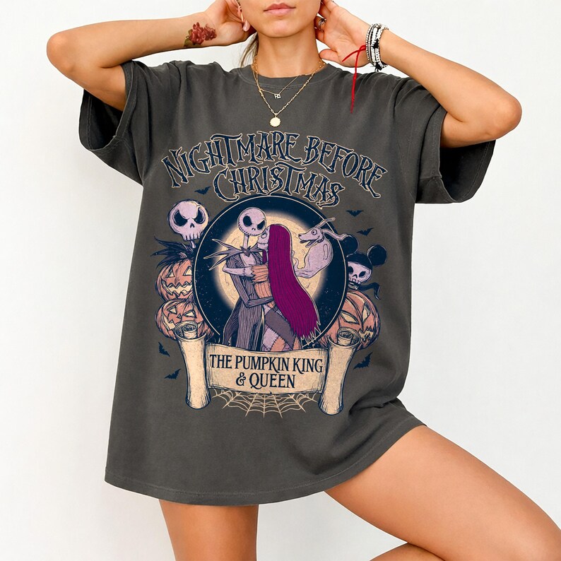 May include: Dark grey t-shirt with a graphic from "The Nightmare Before Christmas." The design shows Jack and Sally embracing, with pumpkins, skulls, and bats. Text reads "Nightmare Before Christmas" and "The Pumpkin King & Queen."