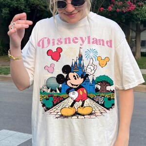 Puede incluir: Camiseta color crema con un gr&aacute;fico de Disneyland. El dise&ntilde;o incluye a Mickey Mouse, un castillo, fuegos artificiales y el texto "Disneyland" en rosa. La camiseta tiene un corte holgado.