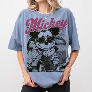 May include: A light blue t-shirt featuring a graphic of Mickey Mouse wearing sunglasses and leaning against a vintage car. The word "Mickey" is printed above the graphic in a distressed, pink font. The shirt has short sleeves and a relaxed fit.
