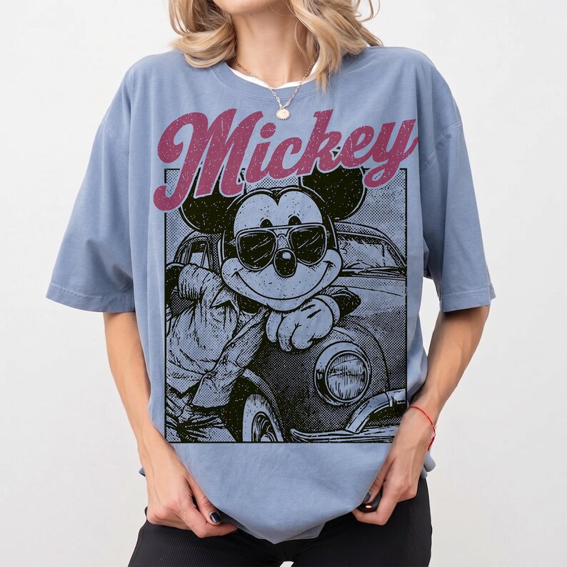 May include: A light blue t-shirt featuring a graphic of Mickey Mouse wearing sunglasses and leaning against a vintage car. The word "Mickey" is printed above the graphic in a distressed, pink font. The shirt has short sleeves and a relaxed fit.