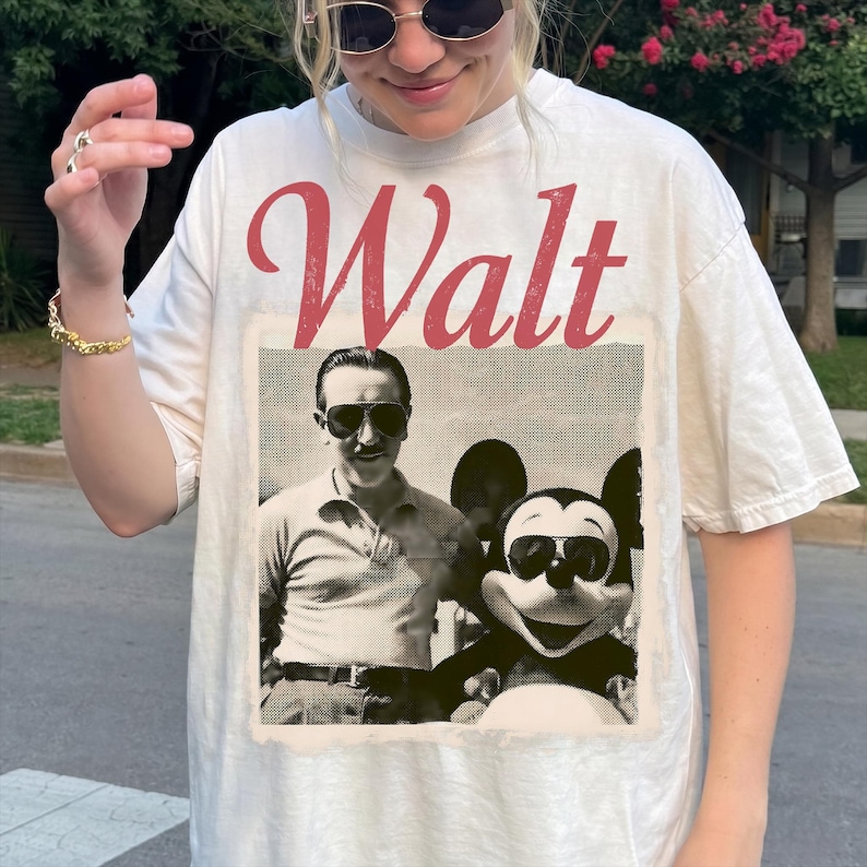 May include: Cream-coloured t-shirt with a vintage graphic of Walt Disney and Mickey Mouse in sunglasses. The word "Walt" is printed in red script. The shirt has a relaxed fit and is made of a soft material.