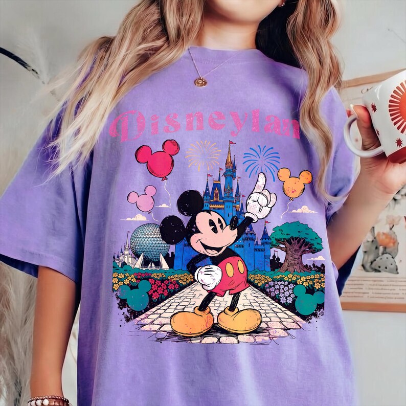 Puede incluir: Una camiseta lila con un gr&aacute;fico de Mickey Mouse se&ntilde;alando un castillo, con la palabra "Disneyland" encima. El dise&ntilde;o incluye globos, fuegos artificiales y detalles florales. La camiseta tiene un corte holgado.