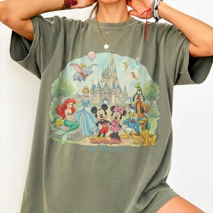 May include: Olive green t-shirt featuring a colorful graphic of Disney characters in front of a castle. The graphic includes Mickey and Minnie Mouse, Ariel, Dumbo, and other characters. The text "Magic Kingdom" and "Walt Disney World" are also visible.