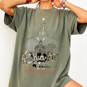 K&ouml;nnte beinhalten: Olivgr&uuml;nes T-Shirt mit einer Grafik von Disney-Figuren und Wahrzeichen, darunter Micky Maus, Minnie Maus und Goofy. Das Design umfasst ein Schloss, einen Baum und den Text "Disneyland Florida".