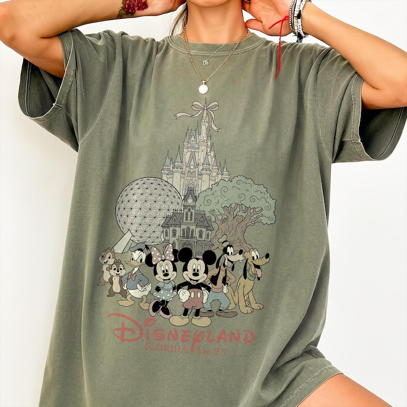 K&ouml;nnte beinhalten: Olivgr&uuml;nes T-Shirt mit einer Grafik von Disney-Figuren und Wahrzeichen, darunter Micky Maus, Minnie Maus und Goofy. Das Design umfasst ein Schloss, einen Baum und den Text "Disneyland Florida".
