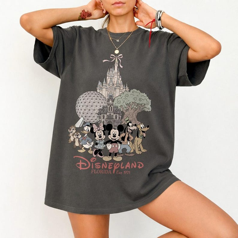 K&ouml;nnte beinhalten: Dunkelgraues T-Shirt mit einem Disneyland Florida-Motiv. Das Design zeigt Micky Maus, Minnie Maus, Donald Duck, Goofy und Pluto, sowie ein Schloss, Spaceship Earth und einen Baum. Der Text "Disneyland Florida Est. 1971" ist darunter gedruckt.