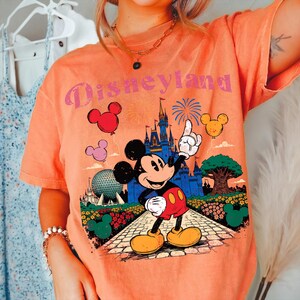 Puede incluir: Camiseta naranja con un gr&aacute;fico de Disneyland que presenta a Mickey Mouse se&ntilde;alando un castillo. El dise&ntilde;o incluye globos, fuegos artificiales y un &aacute;rbol. El texto "Disneyland" es rosa.
