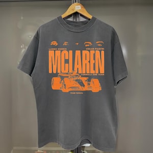 May include: Gray t-shirt featuring the McLaren Formula One Team logo in orange. The shirt includes the names Lando Norris and Oscar Piastri above the McLaren logo, and the words "Formula One Team" and "Team Papaya" below the logo.