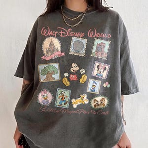 May include: Dark gray t-shirt featuring a Walt Disney World design. The design includes stamps with images of Mickey Mouse, Minnie Mouse, Donald Duck, Goofy, and Chip and Dale. The text "Walt Disney World" and "The Most Magical Place On Earth" are also included.