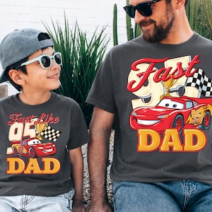 May include: Two matching charcoal gray t-shirts. One shirt has the text "Fast Like 05 DAD" with a red race car graphic, a trophy, and a checkered flag. The other shirt has the text "Fast Dad" with a red race car graphic, a trophy, and a checkered flag.