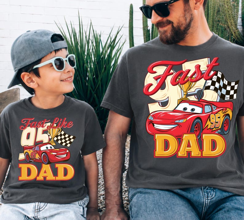 May include: Two matching charcoal gray t-shirts. One shirt has the text "Fast Like 05 DAD" with a red race car graphic, a trophy, and a checkered flag. The other shirt has the text "Fast Dad" with a red race car graphic, a trophy, and a checkered flag.