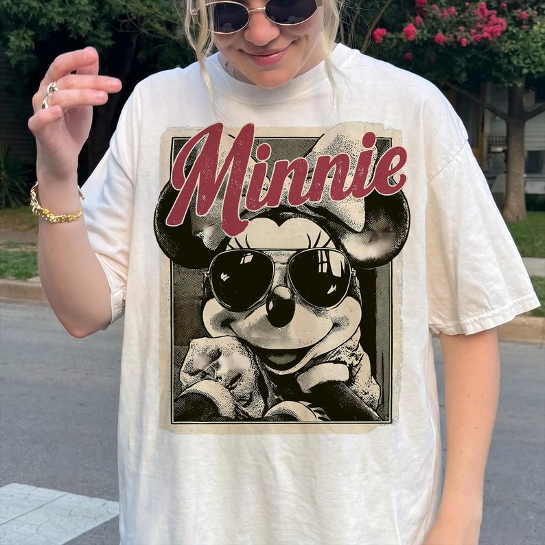 May include: Cream-colored t-shirt featuring a graphic of Minnie Mouse wearing sunglasses, with the name "Minnie" in a cursive font. The image is in a vintage style. The shirt is a casual wear item.