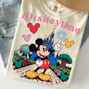 Puede incluir: Camiseta color crema con un gr&aacute;fico de Disneyland que presenta a Mickey Mouse, un castillo y fuegos artificiales. El dise&ntilde;o incluye texto rosa y globos con forma de Mickey Mouse. La camiseta est&aacute; doblada sobre una superficie de mezclilla azul claro.