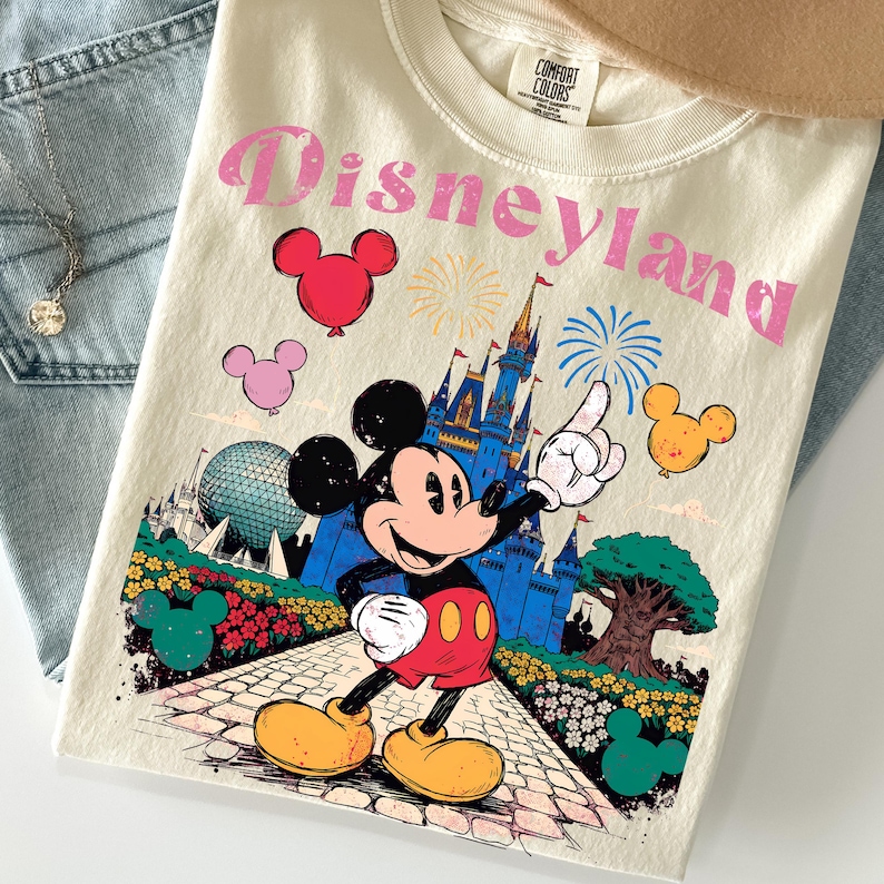 Puede incluir: Camiseta color crema con un gr&aacute;fico de Disneyland que presenta a Mickey Mouse, un castillo y fuegos artificiales. El dise&ntilde;o incluye texto rosa y globos con forma de Mickey Mouse. La camiseta est&aacute; doblada sobre una superficie de mezclilla azul claro.