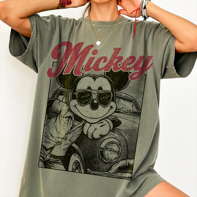 May include: Olive green t-shirt featuring a graphic of Mickey Mouse wearing sunglasses and leaning on a vintage car. The word "Mickey" is printed in a distressed red font across the top of the graphic.