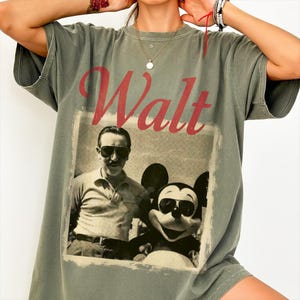 May include: An olive green t-shirt with a vintage graphic of Walt Disney and Mickey Mouse, both wearing sunglasses. The word "Walt" is printed in red script. The shirt has short sleeves and a relaxed fit, suitable for casual wear.
