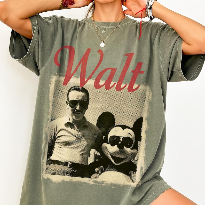 May include: An olive green t-shirt with a vintage graphic of Walt Disney and Mickey Mouse, both wearing sunglasses. The word "Walt" is printed in red script. The shirt has short sleeves and a relaxed fit, suitable for casual wear.