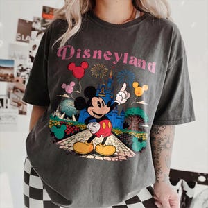 Puede incluir: Camiseta gris oscuro con un gr&aacute;fico colorido de Mickey Mouse se&ntilde;alando un castillo, fuegos artificiales y globos. La palabra "Disneyland" est&aacute; impresa encima del gr&aacute;fico en letras rosas. La camiseta tiene mangas cortas.